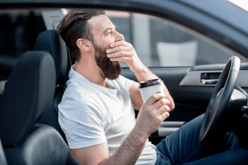 A man yawning in his car while holding a cup of coffee.
